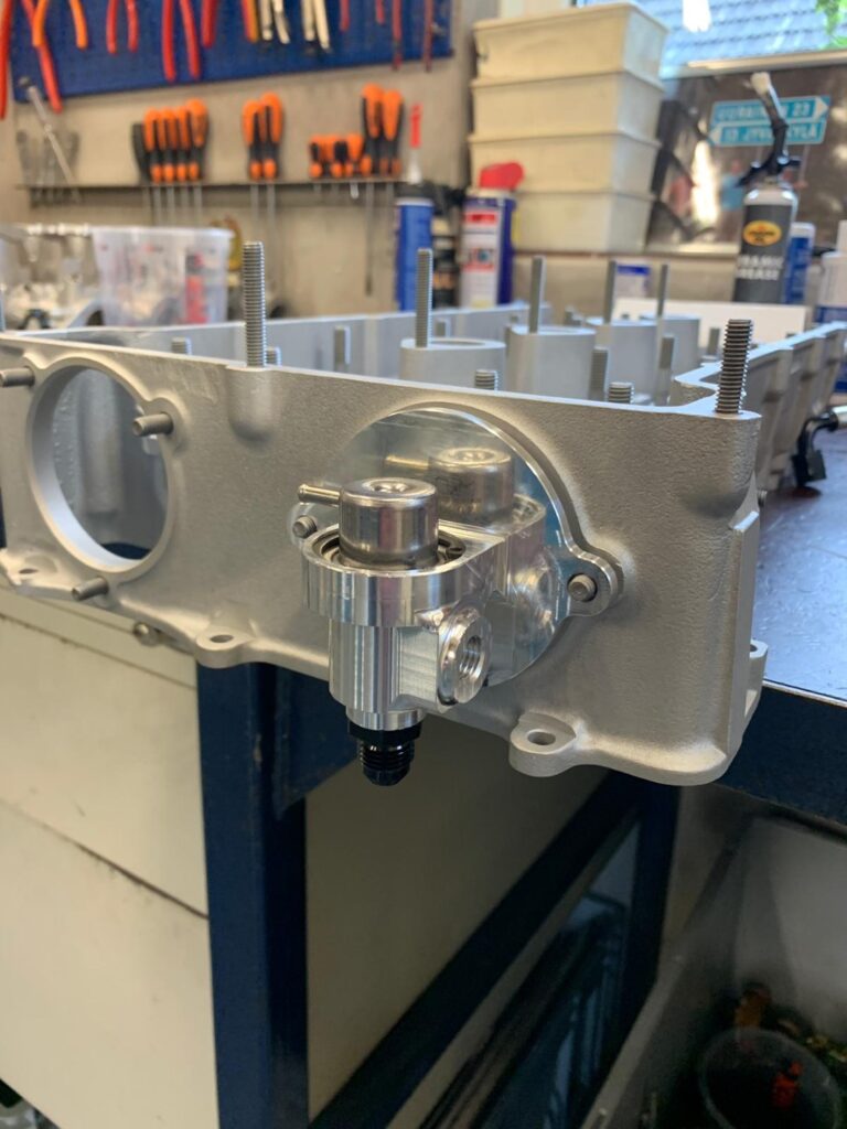 Geassembleerde benzinedrukhouder gemonteerd op een motorblok in werkplaatsomgeving met gereedschap op de achtergrond