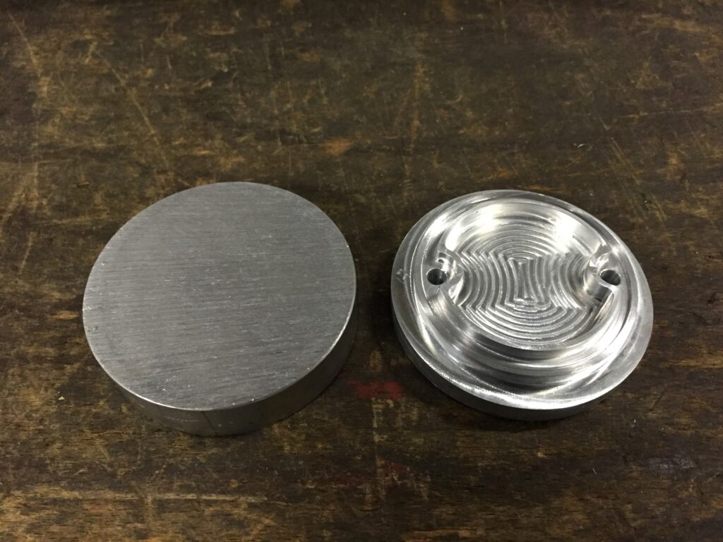 Twee ronde aluminium schijven op een werkbank: links een onbewerkte ruwe schijf, rechts een CNC-bewerkte afdekkap met concentrische freeslijnen en twee montagegaten.