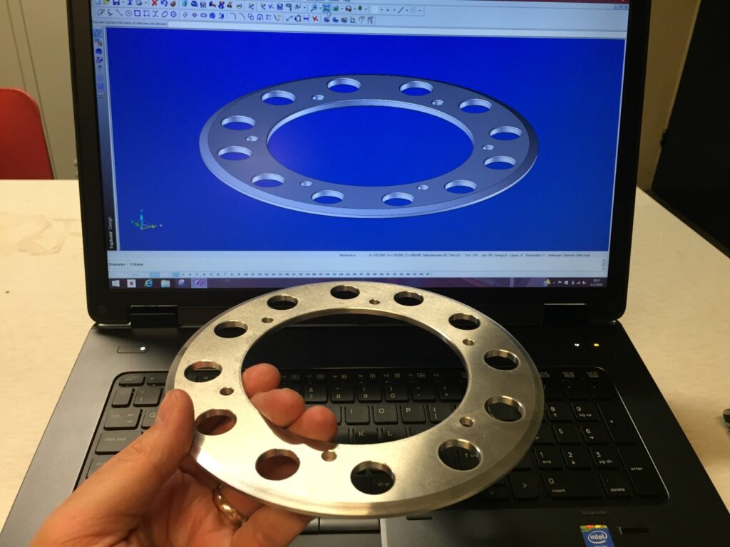 Een hand houdt een gefreesde aluminium adapterring vast voor een laptop waarop het bijbehorende 3D CAD-model wordt weergegeven. De ring en het scherm tonen exact dezelfde geometrie met meerdere gaten in een cirkelpatroon.