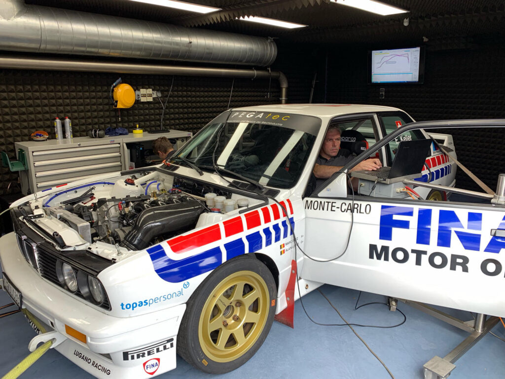 BMW E30 raceauto in FINA-livery tijdens dyno-test met open motorkap en motormanagementanalyse